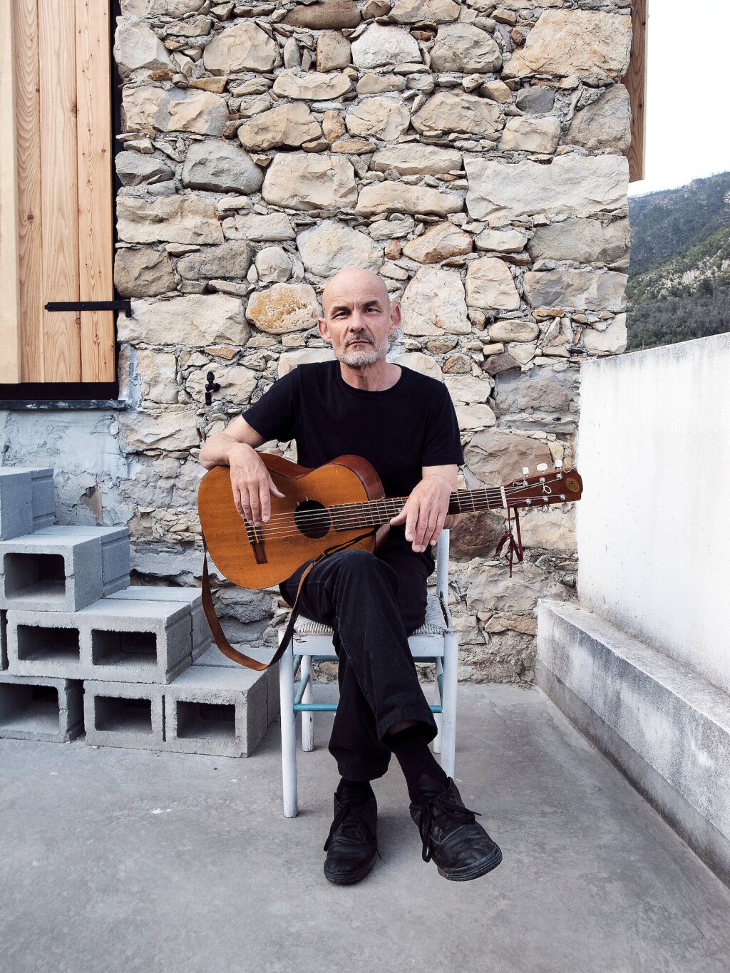 A man with a guitar sits in front of a stone house.