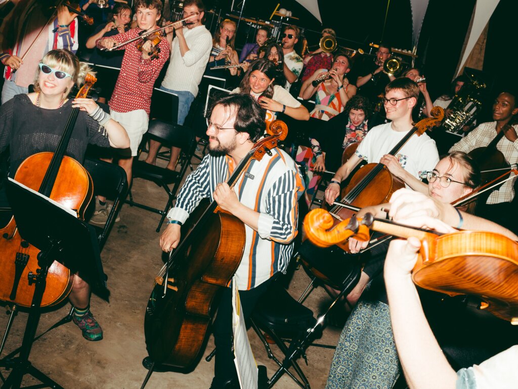 Shot of an orchestra but everyone is dressed in colorful clothing, smiling and there's a lot of movement.