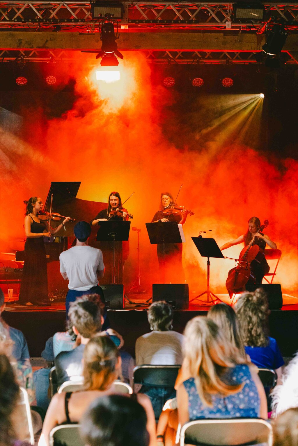 String quartet performing on stage with dramatic orange lights and smoke effects. Audience watching in seated rows.