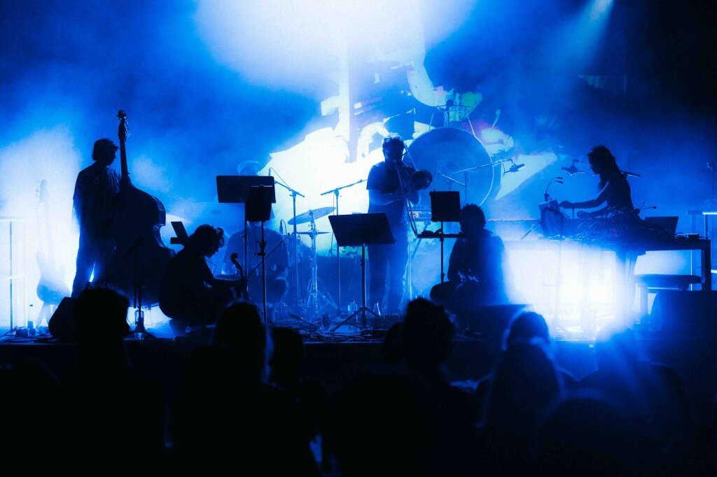 A band with electronic and acoustic instruments on a blue-lit stage.