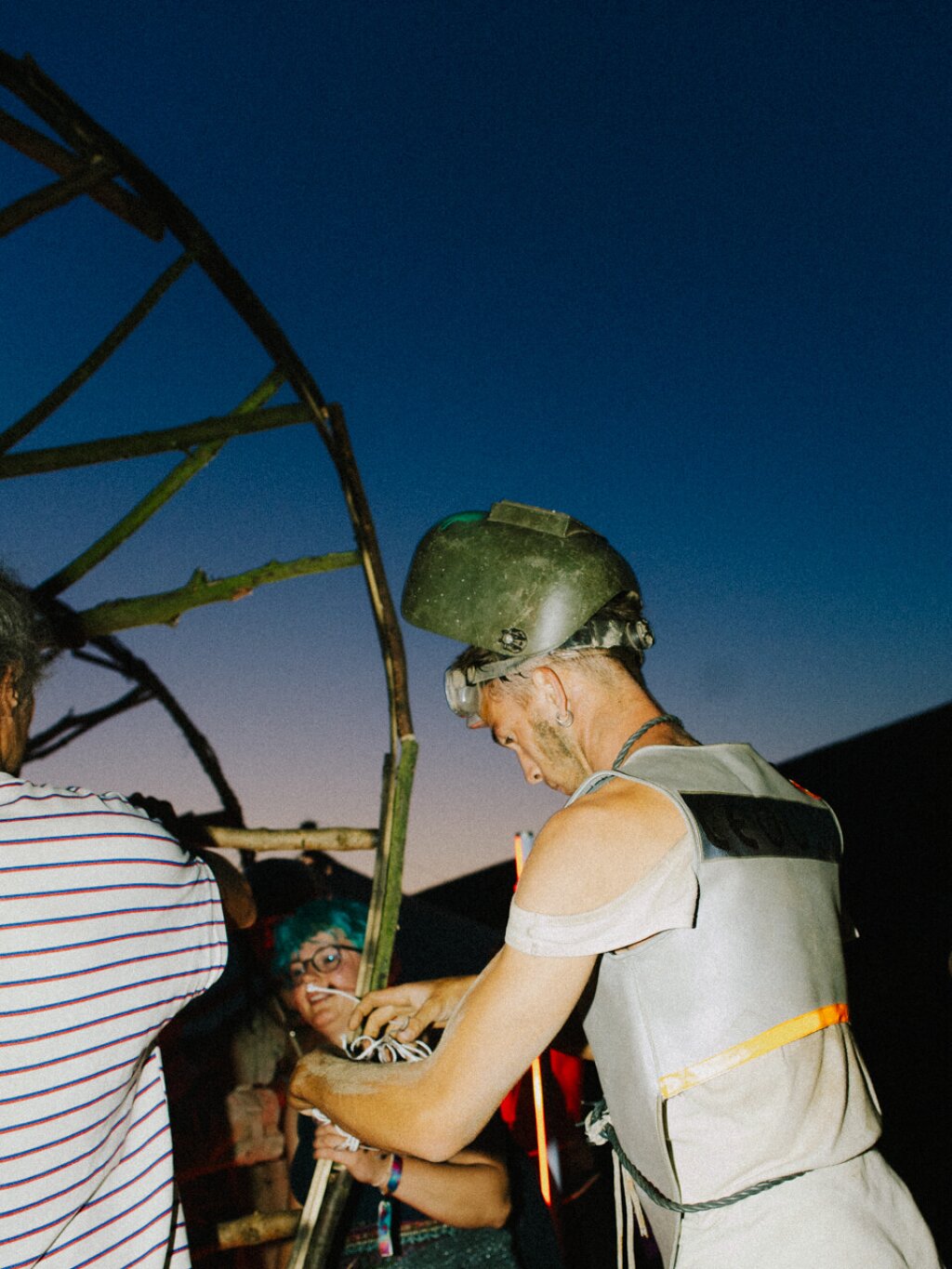 Person in protective gear works on a large wooden structure at dusk, while other people observe.